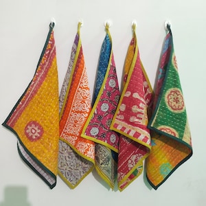 May include: Five colorful, patterned fabric towels hanging on a white wall. The towels feature various designs, including floral and geometric patterns, in shades of yellow, orange, blue, and red. Each towel is trimmed with a contrasting color.
