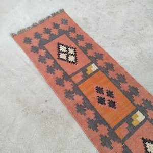 May include: A rectangular runner rug with a geometric pattern. The rug features a salmon-colored background with a central orange rectangle and gray and black geometric shapes. The rug has a fringed edge.