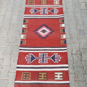 Könnte beinhalten: Ein rechteckiger, handgewebter Teppich mit rotem Hintergrund und geometrischen Mustern in Weiß, Marineblau und Gold. Der Teppich hat ein zentrales Rautenmuster und Fransen. Die Maße betragen ca. 60 cm breit und 122 cm lang.
