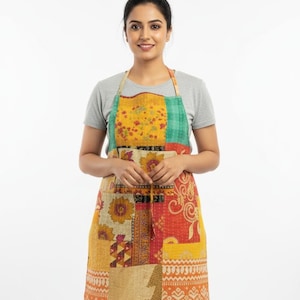 Könnte beinhalten: Ein Patchwork-Schürze mit einer Vielzahl von Mustern und Farben, darunter Gelb, Orange und Grün. Die Schürze hat ein Lätzchen, einen Taillenriemen und eine große Tasche. Die Schürze ist aus einem weichen, gesteppten Stoff gefertigt.