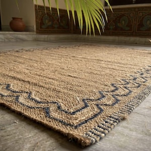 Peut inclure: Un tapis rectangulaire en jute avec une texture tissée et une bordure décorative. Le tapis présente une couleur beige naturel avec un motif géométrique bleu foncé sur les bords. Un article décoratif pour un salon ou une chambre.