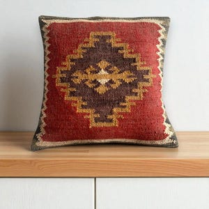 Könnte beinhalten: Quadratisches Zierkissen mit geometrischem Muster. Es hat einen roten Hintergrund mit einem zentralen Rautenmuster in Braun und Gold. Die Ränder sind mit einem dunkelgrauen Rand versehen.
