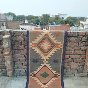 May include: A rectangular rug with a geometric design, featuring diamond shapes in shades of orange, green, and pink against a gray background. The rug has a dark blue border and is displayed outdoors on a brick wall.