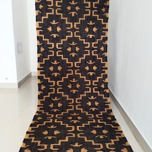 May include: A long, rectangular rug with a geometric pattern. The rug features a repeating design of tan-colored shapes against a dark brown background. The rug is displayed on a white floor and against a white wall.