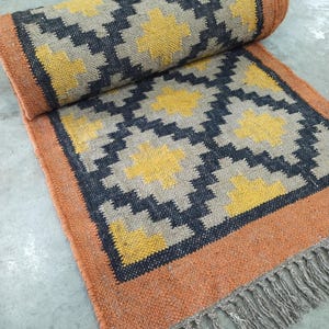 May include: A rolled jute rug with a geometric pattern in yellow, black, and tan. The rug has an orange border and fringe on one end. The design features repeating diamond shapes and starbursts.