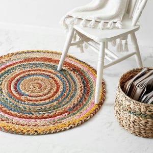 Op de afbeelding: Een rond, gevlochten kleed met concentrische cirkels van veelkleurige stoffen stroken in tinten oranje, blauw, geel en roze, geweven met natuurlijke beige vezels. Het kleed ligt bij een witte stoel en een mand gevuld met tijdschriften.