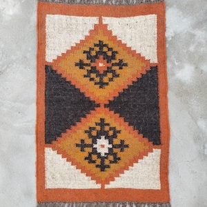 May include: Rectangular jute rug with a geometric diamond pattern in shades of orange, black, and cream. The rug has a fringed edge and a border in a darker orange hue. This rug is suitable for a living room or bedroom.