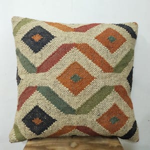 Könnte beinhalten: Quadratisches Zierkissen mit einem geometrischen Rautenmuster in Orange, Grün, Rot und Schwarz auf beigem Hintergrund. Das Kissen besteht aus einem gewebten Stoff und liegt auf einer Holzoberfläche.