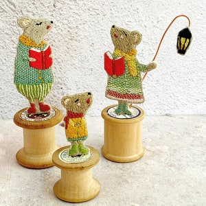 May include: Three embroidered mouse figurines, each holding a book, stand on wooden spools. The mouse on the left is wearing a green and white striped sweater, the middle mouse is wearing a green dress with a yellow scarf, and the mouse on the right is holding a lantern.