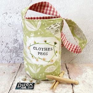 May include: A green fabric bag with a red and white checkered handle. The bag has a white label with the text "Clothes Pegs" embroidered on it. The bag is sitting on a wooden surface with wooden clothespins.