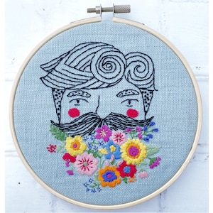 Puede incluir: Arte de bordado en aro con un rostro estilizado con bigote y un ramo floral colorido. El rostro tiene un fondo azul y contornos negros. Las flores son rosas, amarillas, naranjas, azules y verdes.