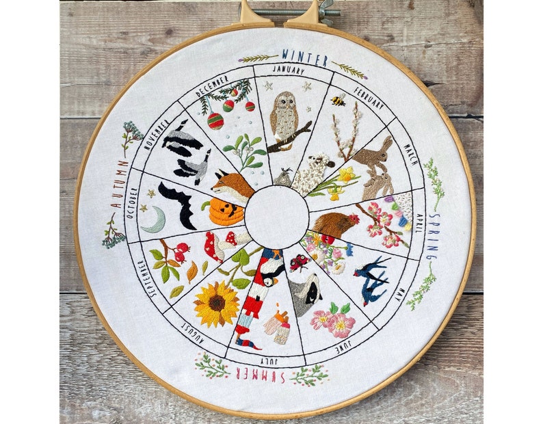 May include: Embroidered hoop art depicting a circular calendar with each month represented by a different animal and floral design. The months are labeled around the circle, with the seasons "Winter", "Spring", "Summer", and "Autumn" in larger text.
