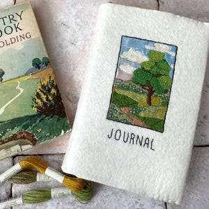 May include: A white journal with a hand-stitched embroidered landscape scene of a green tree and a winding path. The journal cover reads "JOURNAL".