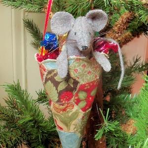May include: A grey felt mouse holding a cone-shaped fabric stocking filled with wrapped sweets. The stocking is decorated with a patchwork of floral and geometric patterns in shades of brown, green, red, and blue.