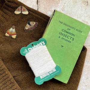 May include: A brown sweater with three embroidered moths. A spool of white thread is on top of a green book titled "The Observer's Book of Common Insects & Spiders".