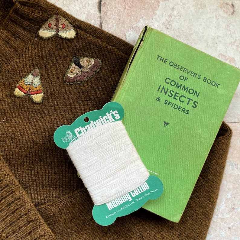 May include: A brown sweater with three embroidered moths. A spool of white thread is on top of a green book titled "The Observer's Book of Common Insects & Spiders".