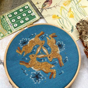 Pu&ograve; includere: Ricamo su telaio con tre lepri marroni su sfondo blu-verde, circondate da motivi di dente di leone bianchi. Il telaio in legno misura circa 20 cm di diametro. Una scheda di bottoni vintage e una stampa botanica sono sullo sfondo.