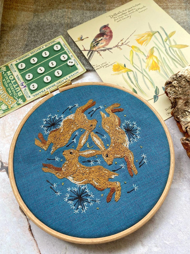 Pu&ograve; includere: Ricamo su telaio con tre lepri marroni su sfondo blu-verde, circondate da motivi di dente di leone bianchi. Il telaio in legno misura circa 20 cm di diametro. Una scheda di bottoni vintage e una stampa botanica sono sullo sfondo.