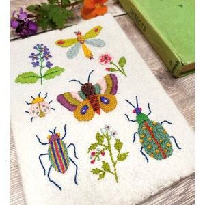 May include: Embroidered white fabric featuring colorful insect and floral designs. The artwork includes a dragonfly, butterfly, beetles, and flowers, all meticulously stitched with various colors. A green book titled "Insects" is visible in the background.