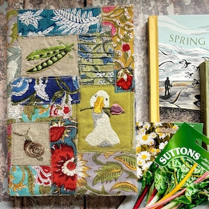 May include: A rectangular fabric-covered journal featuring a patchwork design of floral and botanical prints in blue, red, green, and yellow. The cover includes embroidered details of a pea pod, a snail, and a white duck holding a purple flower.