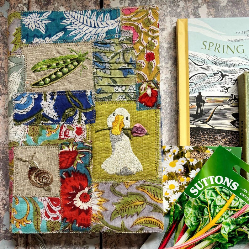 May include: A rectangular fabric-covered journal featuring a patchwork design of floral and botanical prints in blue, red, green, and yellow. The cover includes embroidered details of a pea pod, a snail, and a white duck holding a purple flower.