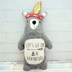 May include: Gray plush bear wearing a red and yellow headband with a green feather. The bear has a white patch on its chest with the text "Let's go on an adventure" and an arrow pointing to the right.