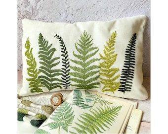 May include: Cream-colored rectangular pillow embroidered with various green fern designs. The ferns vary in shades of green and black. The pillow is placed on a surface with vintage botanical illustrations and spools of thread.