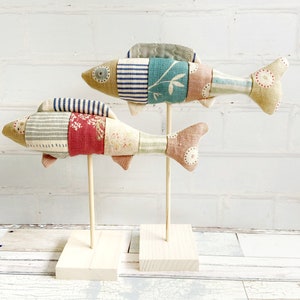 May include: Two patchwork fabric fish on wooden stands. The fish are made of different coloured fabrics with a striped pattern and floral designs. The fish are displayed against a white brick background.