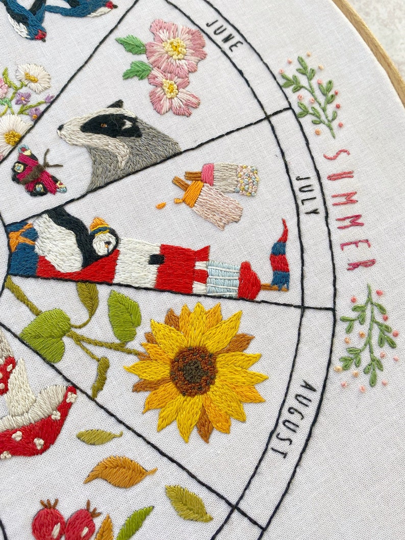 May include: Embroidered hoop art depicting the months of June, July, and August. The design features a badger, ice cream cones, a sunflower, a lighthouse, a puffin, and a red mushroom.