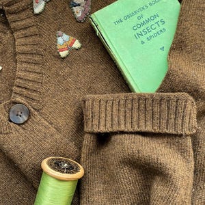 May include: A brown knitted cardigan with a green book titled "The Observer's Book of Common Insects & Spiders" tucked into the pocket. There are three embroidered butterflies on the cardigan, and a spool of green thread is on the bottom left.