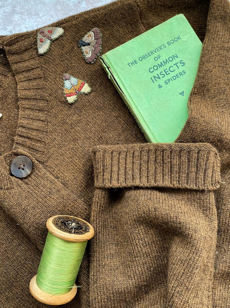 May include: A brown knitted cardigan with a green book titled "The Observer's Book of Common Insects & Spiders" tucked into the pocket. There are three embroidered butterflies on the cardigan, and a spool of green thread is on the bottom left.