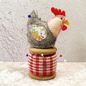 May include: A grey felt pincushion shaped like a chicken with a red comb and yellow beak, sitting on a wooden spool wrapped in red and white gingham fabric. The pincushion has a floral fabric patch on its back and is decorated with colourful pins.