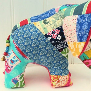 Peut inclure: Un &eacute;l&eacute;phant en peluche fait de morceaux de tissu color&eacute;s. L'&eacute;l&eacute;phant a une clochette &agrave; la queue et se tient sur une surface blanche.