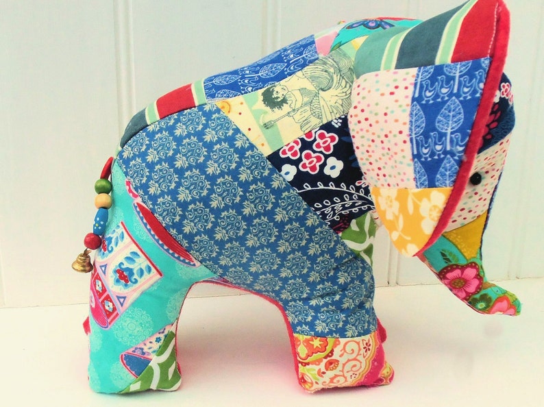 Peut inclure: Un &eacute;l&eacute;phant en peluche fait de morceaux de tissu color&eacute;s. L'&eacute;l&eacute;phant a une clochette &agrave; la queue et se tient sur une surface blanche.