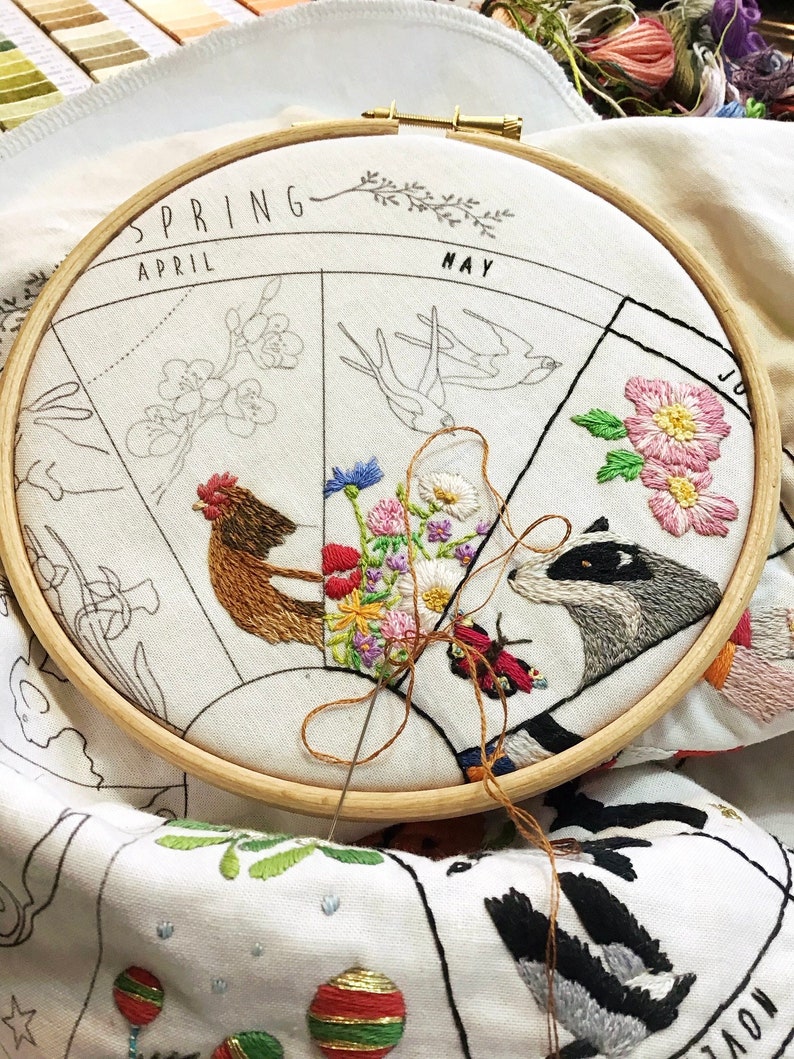 May include: A close-up of a circular embroidery hoop with a white fabric background. The fabric is embroidered with a spring-themed design, including a rooster, flowers, birds, and a raccoon. The hoop is made of wood and has a brown finish. The text "SPRING", "APRIL", and "MAY" is visible on the hoop.