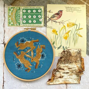 Pu&ograve; includere: Un ricamo con tre lepri marroni in cerchio su sfondo blu-verde. Sono presenti anche una cartolina vintage con un uccello e narcisi, una scheda bottoni e un pezzo di corteccia.