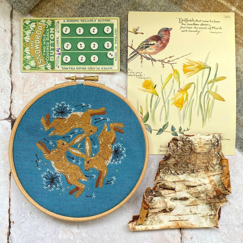 Pu&ograve; includere: Un ricamo con tre lepri marroni in cerchio su sfondo blu-verde. Sono presenti anche una cartolina vintage con un uccello e narcisi, una scheda bottoni e un pezzo di corteccia.