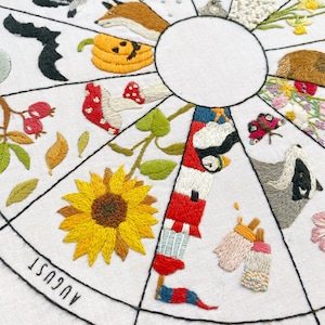 May include: Embroidered white fabric circle with a black outline, divided into 12 sections, each representing a month of the year. Each section features a different embroidered image, such as a sunflower, a pumpkin, a fox, a bat, a bird, a butterfly, and a badger. The months are labeled in black text around the circle.