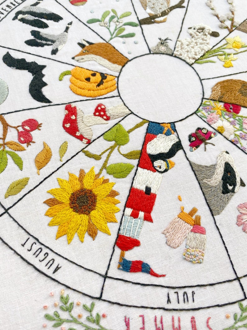 May include: Embroidered white fabric circle with a black outline, divided into 12 sections, each representing a month of the year. Each section features a different embroidered image, such as a sunflower, a pumpkin, a fox, a bat, a bird, a butterfly, and a badger. The months are labeled in black text around the circle.