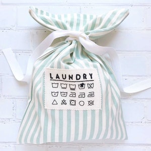Op de afbeelding: Een groen-wit gestreepte waszak met een wit trekkoord. De zak heeft een patch met het woord "LAUNDRY" en verschillende wasverzorgingssymbolen.