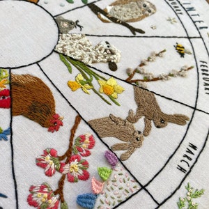 May include: Embroidered hoop art depicting a calendar of the months of the year with various animals and plants. The months are labeled in a circle around the hoop, with January at the top and December at the bottom. The animals and plants are embroidered in different colors, including red, green, blue, yellow, and brown.