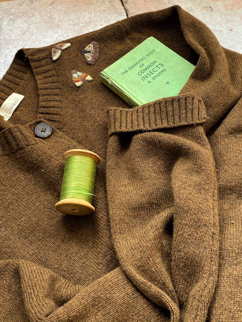 May include: A brown knitted cardigan with a button closure and a small green spool of thread. The cardigan has three embroidered butterflies on the front. A green book titled "The Observer's Book of Common Insects & Spiders" is tucked into the cardigan's pocket.