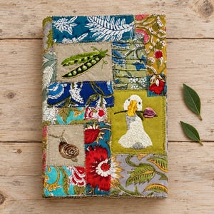 May include: A rectangular fabric cover featuring a patchwork design of various floral and botanical prints in blue, green, red, and yellow. Embroidered details include a green pea pod, a brown snail, and a white goose holding a pink flower.