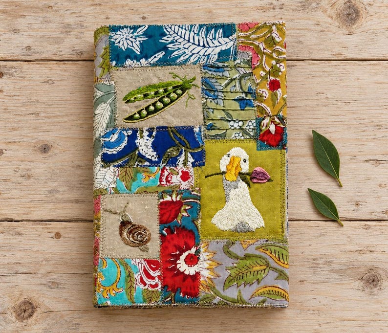 May include: A rectangular fabric cover featuring a patchwork design of various floral and botanical prints in blue, green, red, and yellow. Embroidered details include a green pea pod, a brown snail, and a white goose holding a pink flower.