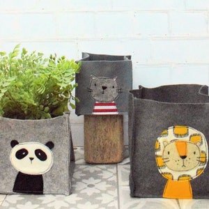 Op de afbeelding: Drie grijze felt opbergbakken met dierengezichten. De bak links heeft een panda gezicht, de middelste bak heeft een kattengezicht en de bak rechts heeft een leeuwengezicht.