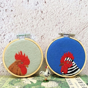 May include: Two embroidered hoop art pieces featuring roosters. One rooster is brown with a red comb and wattle on a light green background. The other rooster is black and white with a red comb and wattle on a blue background.