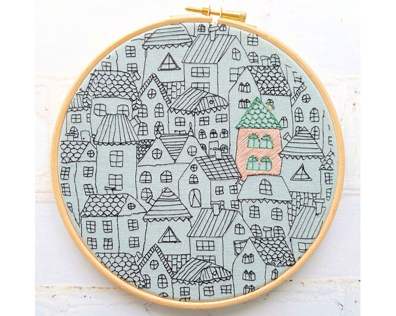 May include: A circular embroidery hoop with a light blue background and black embroidery of a city skyline with houses. The houses are all different shapes and sizes and have windows and doors. The embroidery is detailed and intricate.