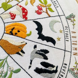 May include: Embroidered hoop art depicting a seasonal calendar with illustrations of autumn and winter months. The months are labeled September, October, November, and December. The illustrations include a sunflower, mushrooms, a pumpkin, a bat, a crescent moon, a fox, geese, mistletoe, and a Christmas ornament.