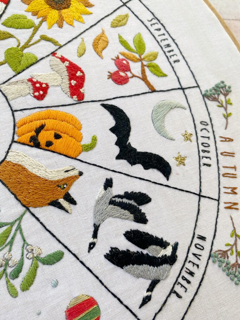 May include: Embroidered hoop art depicting a seasonal calendar with illustrations of autumn and winter months. The months are labeled September, October, November, and December. The illustrations include a sunflower, mushrooms, a pumpkin, a bat, a crescent moon, a fox, geese, mistletoe, and a Christmas ornament.