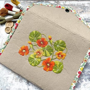 Puede incluir: Estuche de fieltro beige con un diseño floral bordado con flores naranjas y amarillas y hojas verdes. El estuche está adornado con una tela estampada floral. Hilos de bordar y botones visibles.